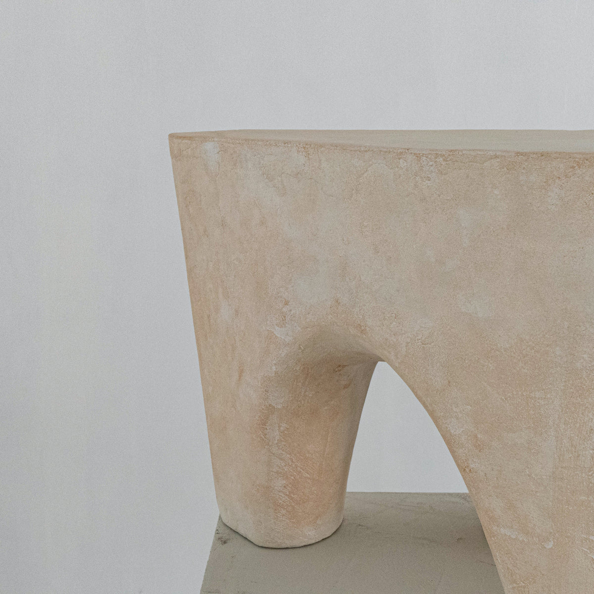 A close-up of a handmade abstract sculptural stool with a soft beige finish, featuring a wide top rim and a central arched opening, displayed on a gray pedestal against a neutral gray background.