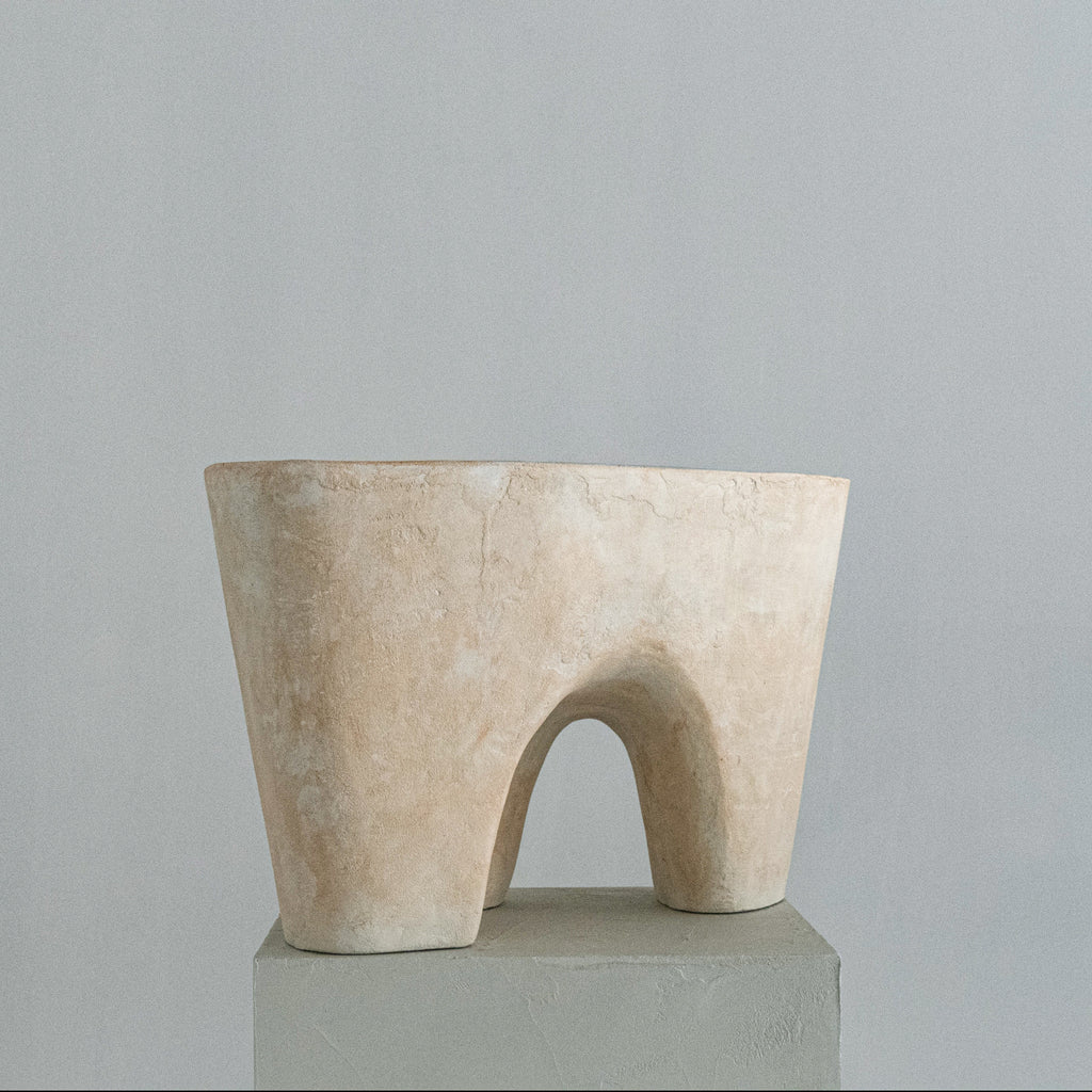A handmade abstract sculptural stool with a soft beige finish, featuring a wide top rim and a central arched opening, displayed on a gray pedestal against a neutral gray background.