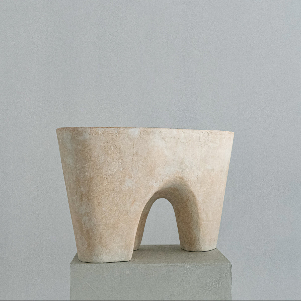 A handmade abstract sculptural stool with a soft beige finish, featuring a wide top rim and a central arched opening, displayed on a gray pedestal against a neutral gray background.