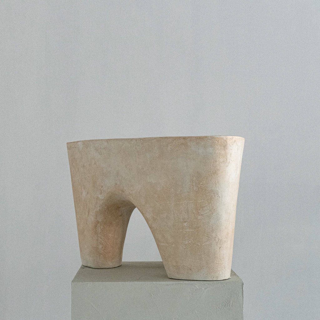 A handmade abstract sculptural stool with a soft beige finish, featuring a wide top rim and a central arched opening, displayed on a gray pedestal against a neutral gray background.