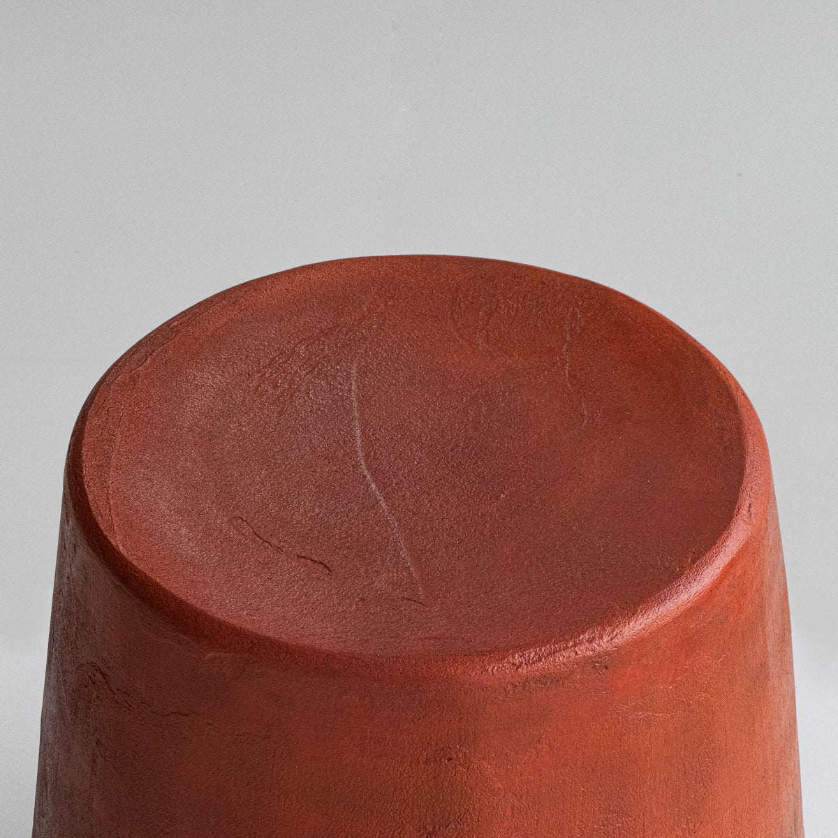 A sculptural stool finished by hand in micro-cement, left deliberately rough and textured rather than smoothed — letting the raw character of the natural stone material show through. The finish carries a subtle color gradient, a warm blood-orange tone at the seat fading gradually into dark grey toward the base, giving each piece its own distinctive, one-of-a-kind character.
