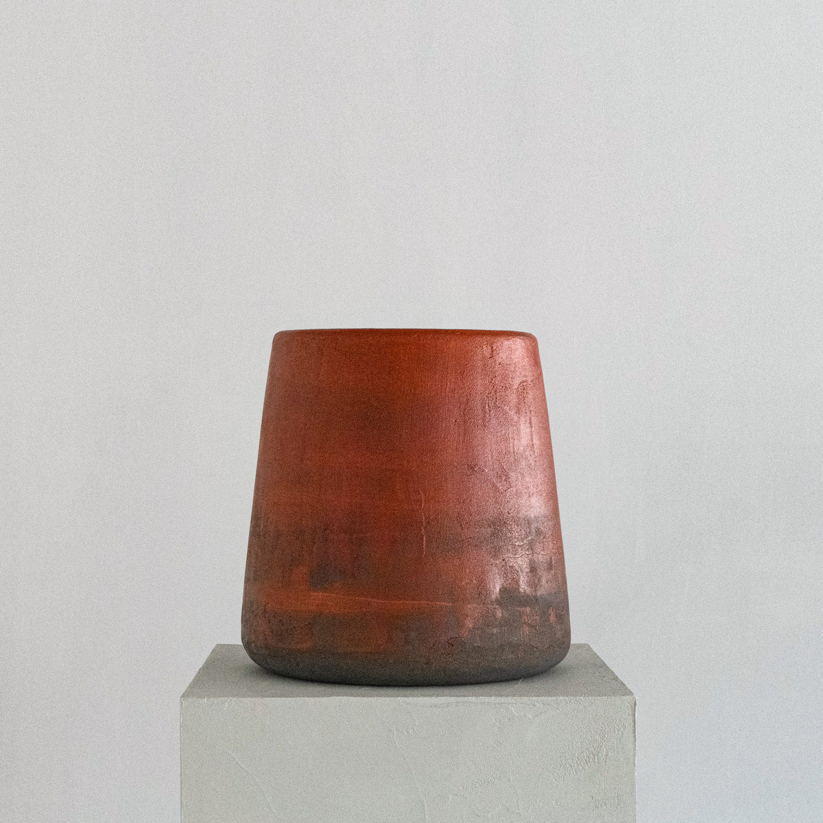 A sculptural stool finished by hand in micro-cement, left deliberately rough and textured rather than smoothed — letting the raw character of the natural stone material show through. The finish carries a subtle color gradient, a warm blood-orange tone at the seat fading gradually into dark grey toward the base, giving each piece its own distinctive, one-of-a-kind character.
