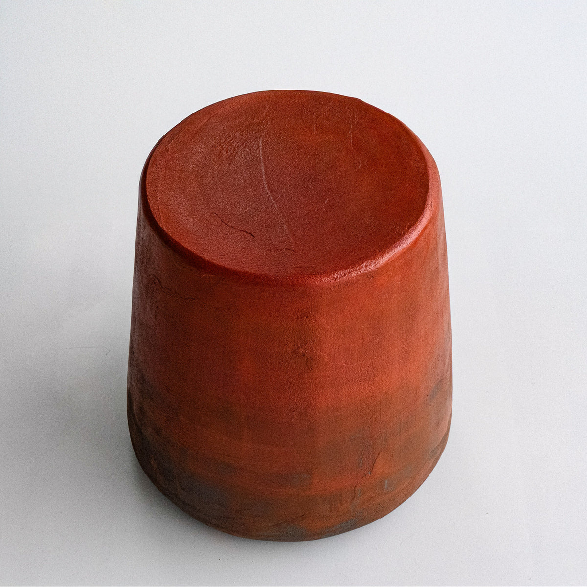 A sculptural stool finished by hand in micro-cement, left deliberately rough and textured rather than smoothed — letting the raw character of the natural stone material show through. The finish carries a subtle color gradient, a warm blood-orange tone at the seat fading gradually into dark grey toward the base, giving each piece its own distinctive, one-of-a-kind character.