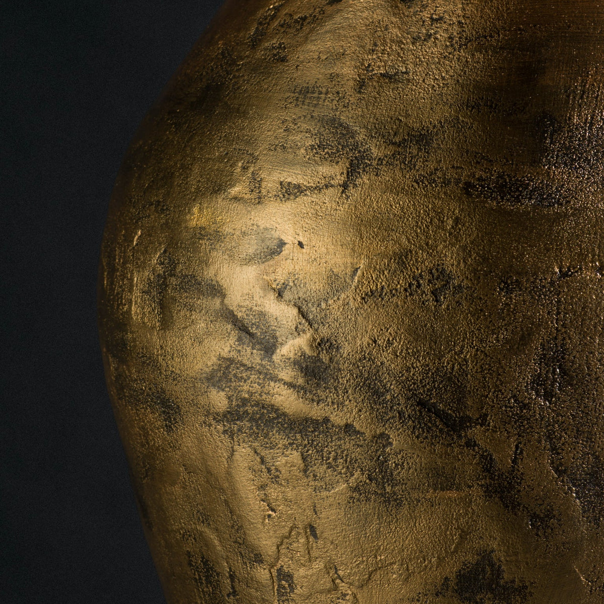 Close-up of a textured metallic gold stool against a dark black background, showing a hammered or brushed finish with tonal variation.