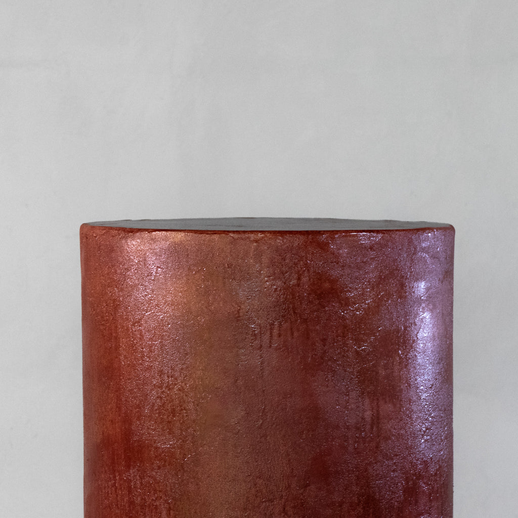A cylindrical micro-cement stool in a bloody orange color, with a rough-textured, glossy finish, displayed on a white pedestal against a neutral grey wall.