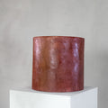 A cylindrical micro-cement stool in a bloody orange color, with a rough-textured, glossy finish, displayed on a white pedestal against a neutral grey wall.