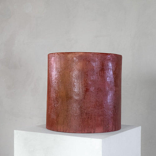 A cylindrical micro-cement stool in a bloody orange color, with a rough-textured, glossy finish, displayed on a white pedestal against a neutral grey wall.
