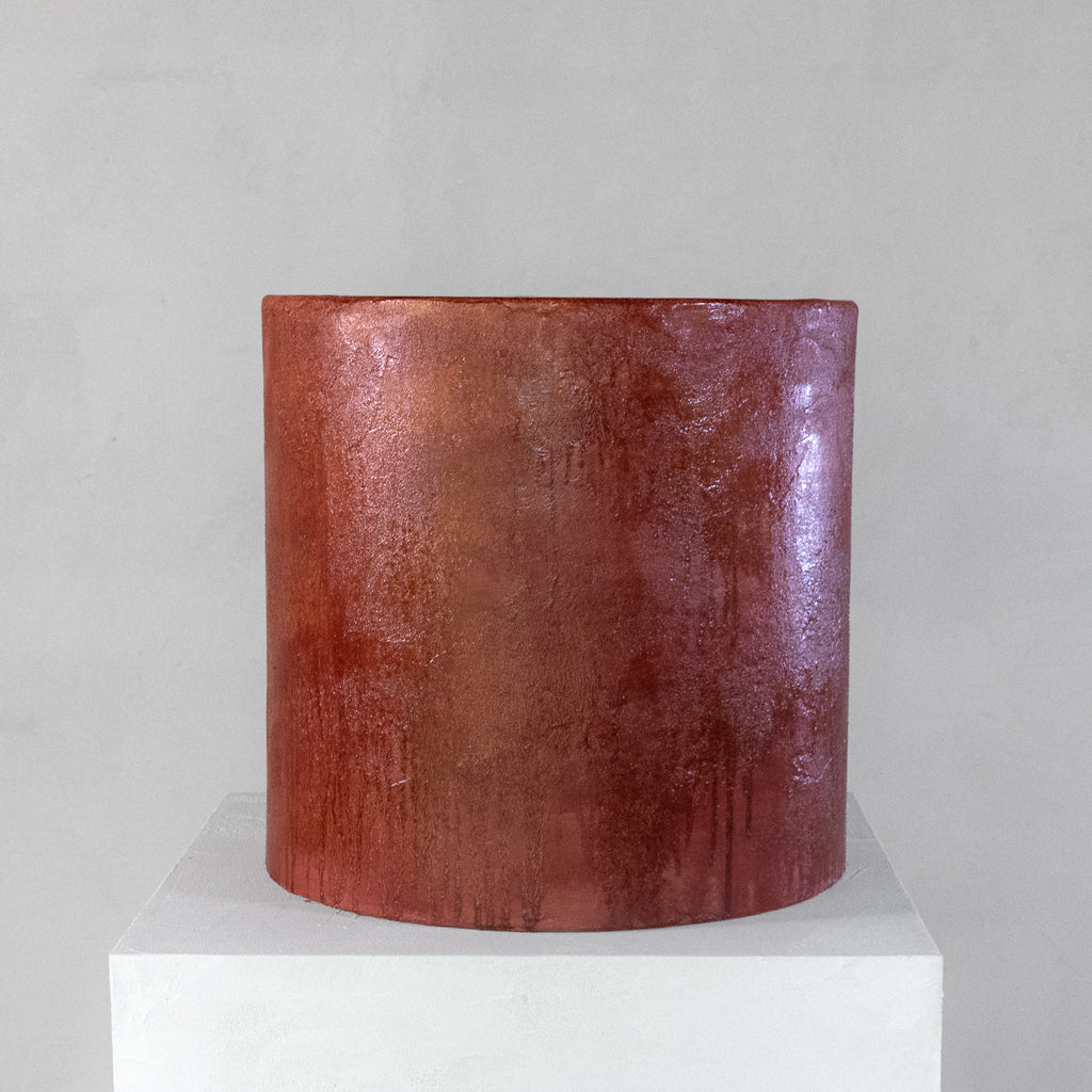 A cylindrical micro-cement stool in a bloody orange color, with a rough-textured, glossy finish, displayed on a white pedestal against a neutral grey wall.