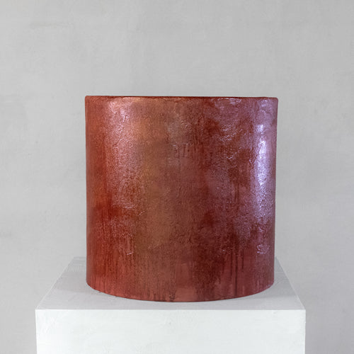 A cylindrical micro-cement stool in a bloody orange color, with a rough-textured, glossy finish, displayed on a white pedestal against a neutral grey wall.