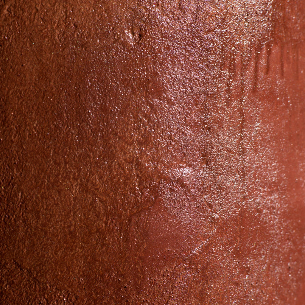 A close-up of a cylindrical micro-cement stool in a bloody orange color, with a rough-textured, glossy finish, displayed on a white pedestal against a neutral grey wall.