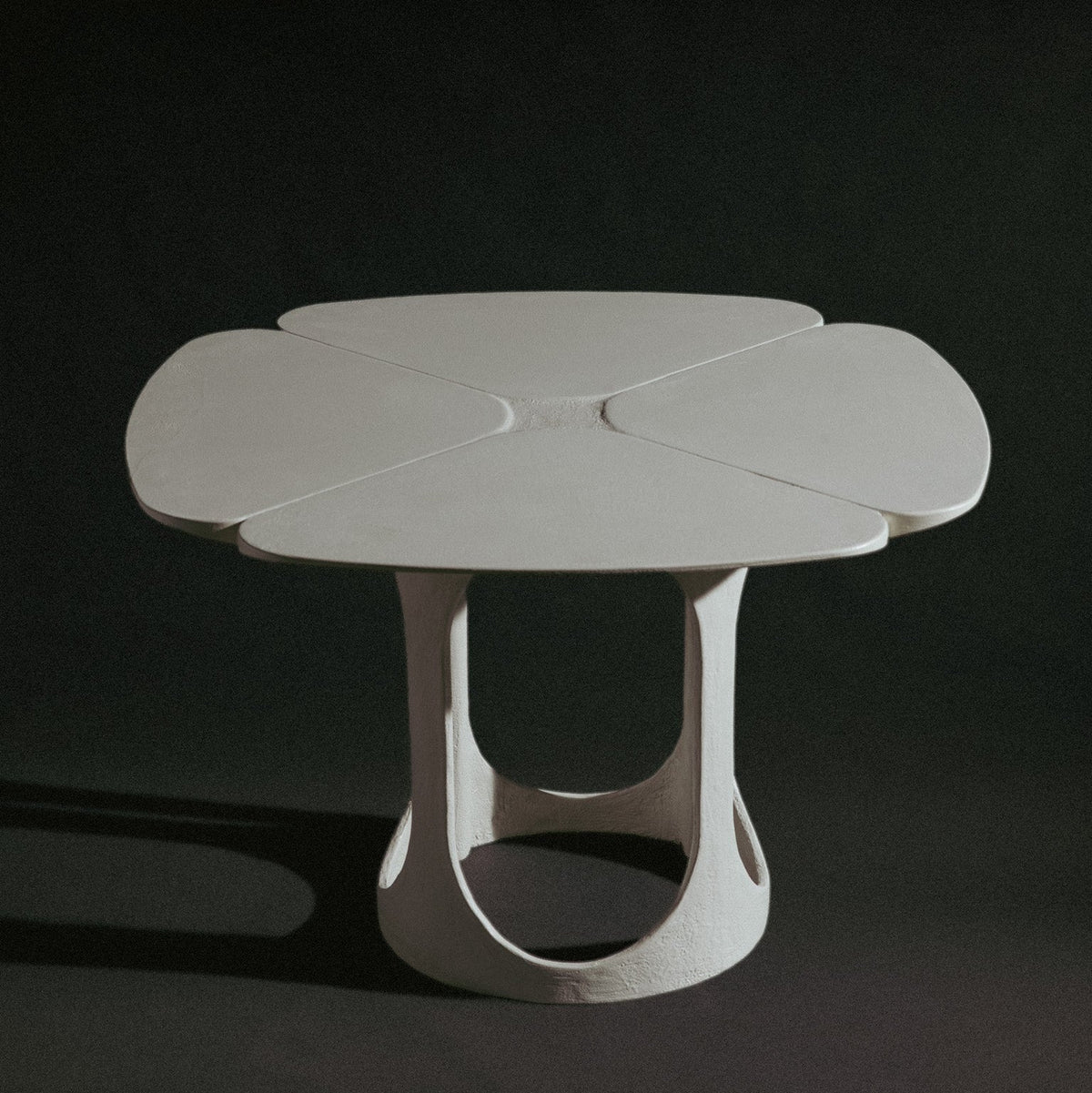 A handmade sculptural, feminine table with a four-petal clover-shaped top and a textured, organic-shaped base, shown on a dark studio background.