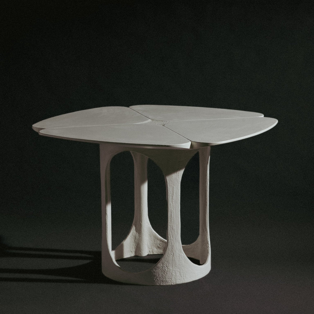 A handmade sculptural, feminine table with a four-petal clover-shaped top and a textured, organic-shaped base, shown on a dark studio background.