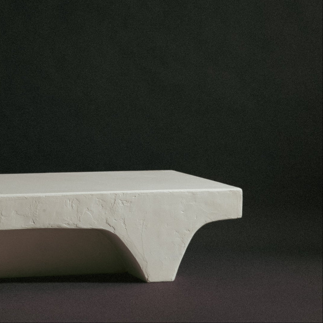 A minimalist rectangular bench with a thick, textured off-white stone or concrete top and sculptural angular legs.