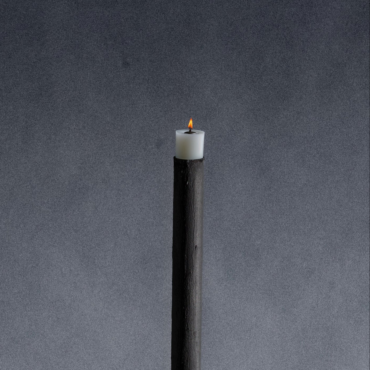A closeup of a minimalist black metal pole candle holder stands vertically on a stone painted in a bright blue base.