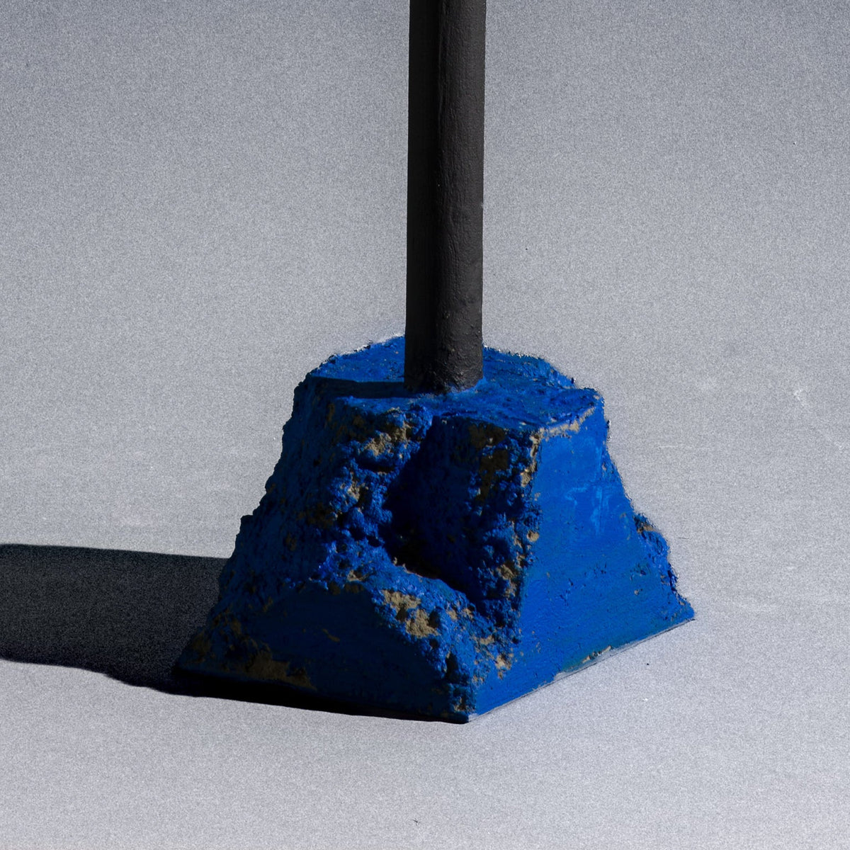 A close-up of a blue textured concrete or stone sculpture with a rough. A minimalist black metal pole candle holder stands vertically on a stone painted in a bright blue base.