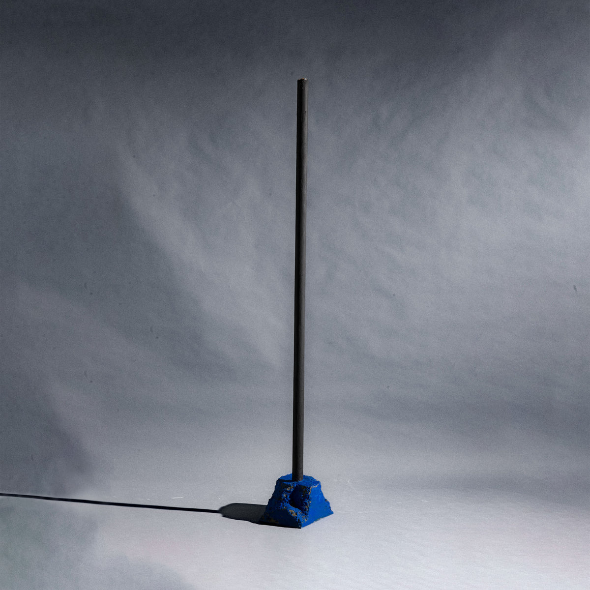 A minimalist black metal pole candle holder stands vertically on a stone painted in a bright blue base.