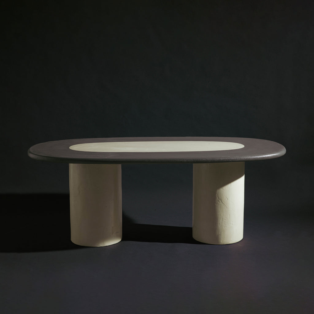 A handmade long oval dining table with a dark gray stone top, a light beige inlay, and two thick cylindrical off-white legs, photographed against a dark studio background.