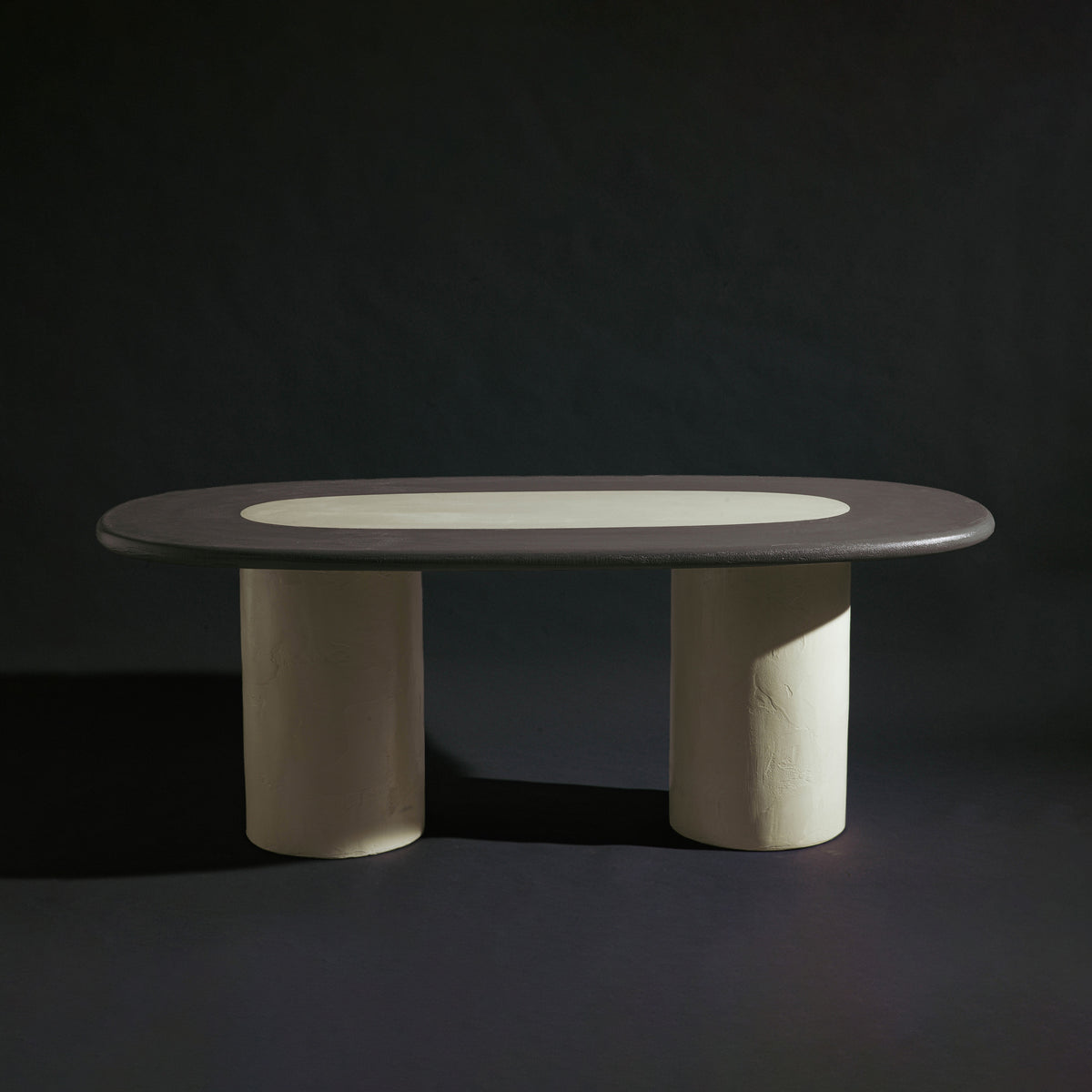 A handmade long oval dining table with a dark gray stone top, a light beige inlay, and two thick cylindrical off-white legs, photographed against a dark studio background.