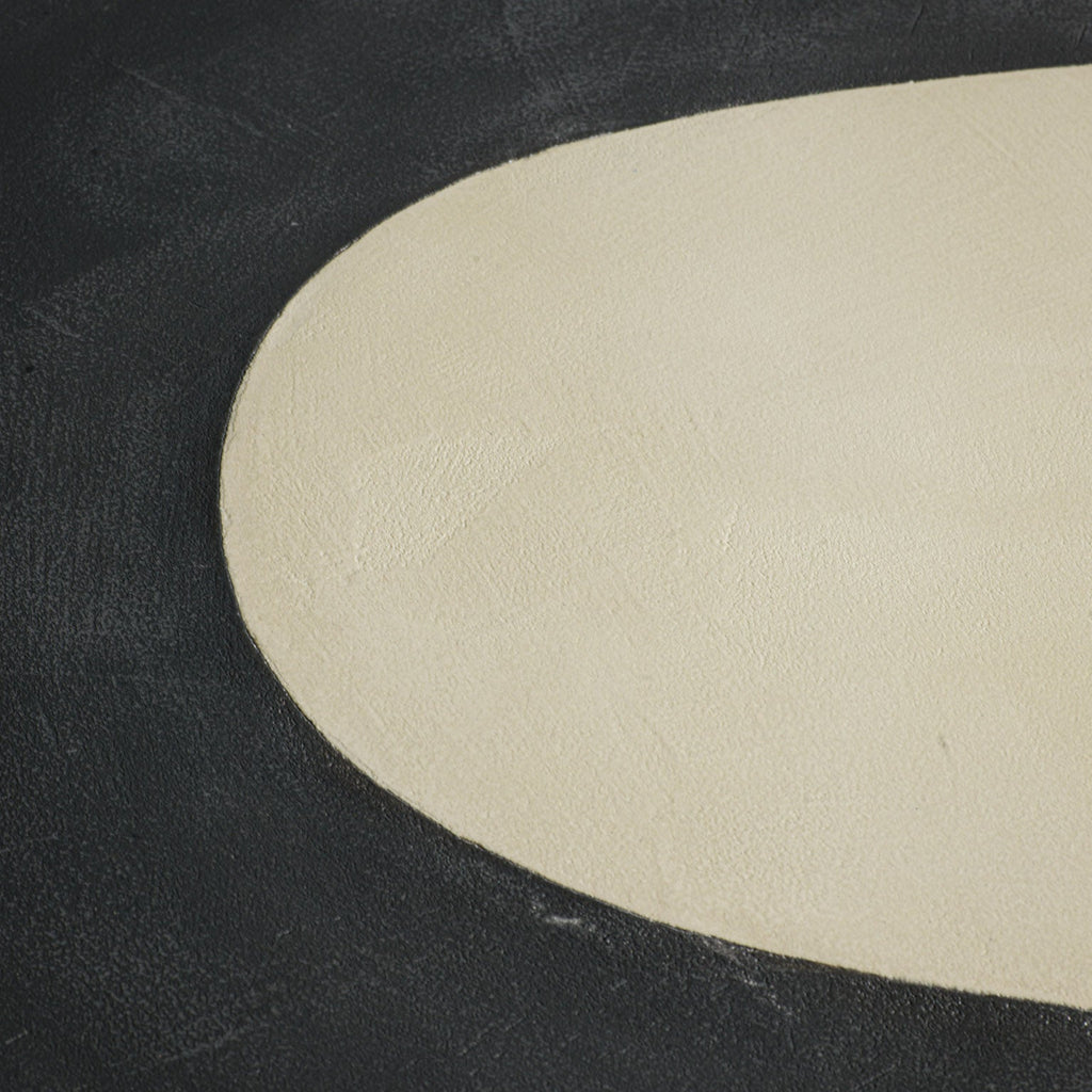 A close up of a handmade long oval dining table with a dark gray stone top, a light beige inlay, and two thick cylindrical off-white legs, photographed against a dark studio background.