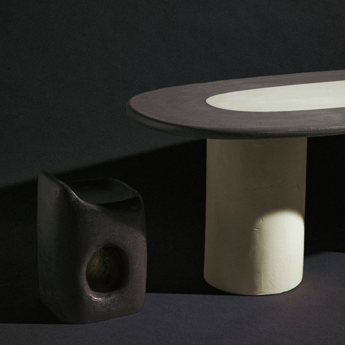 A modern sculptural table with a dark charcoal oval top and a thick cylindrical off-white pedestal base, shown next to a matching dark textured side container on a dark studio background.