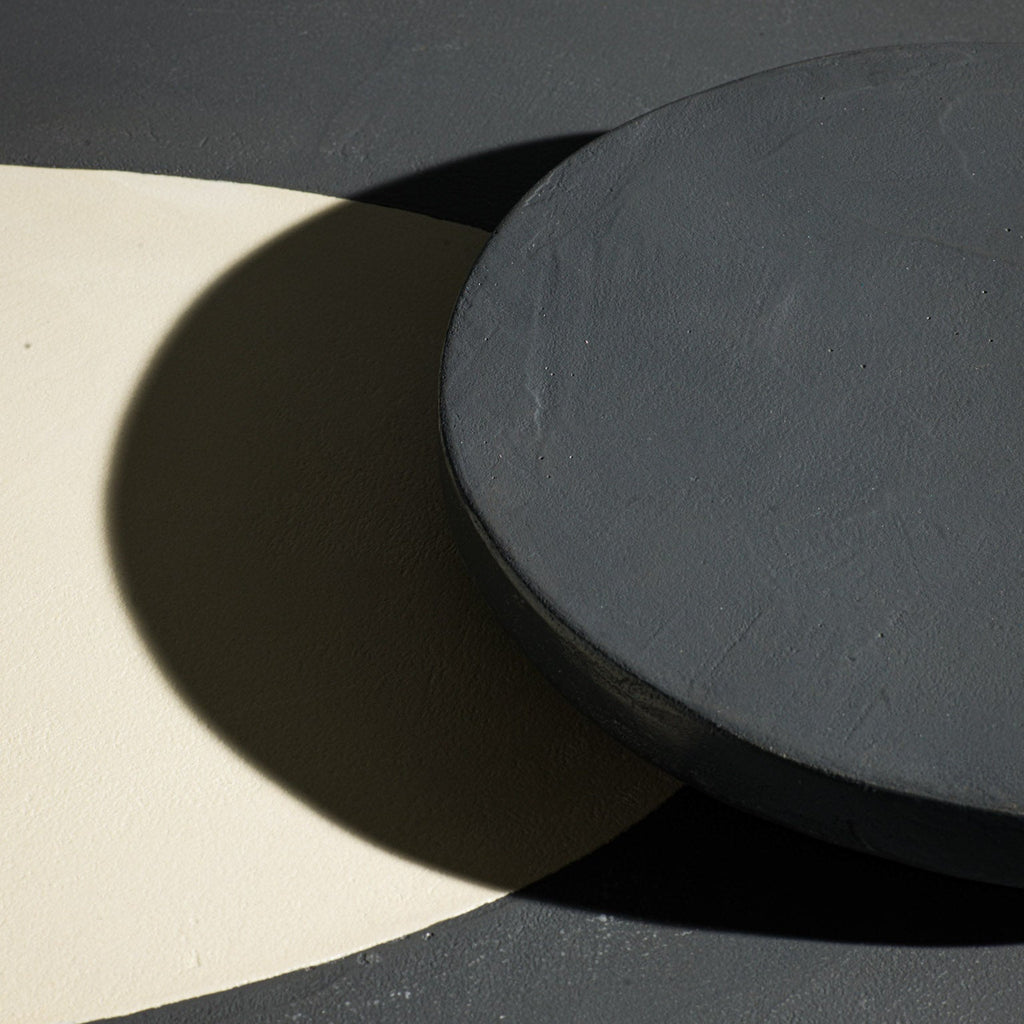 A close-up of a dark charcoal grey round bread plate resting on table with a dark gray and a large cream-colored top.