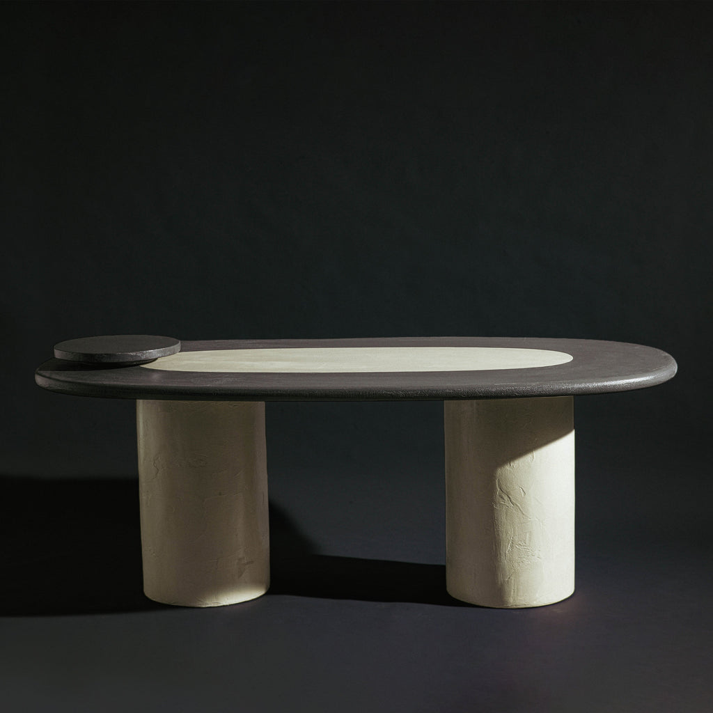 A handmade long oval dining table with a dark gray stone top, a light beige inlay, and two thick cylindrical off-white legs, photographed against a dark studio background.