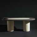 A handmade long oval dining table with a dark gray stone top, a light beige inlay, and two thick cylindrical off-white legs, photographed against a dark studio background.