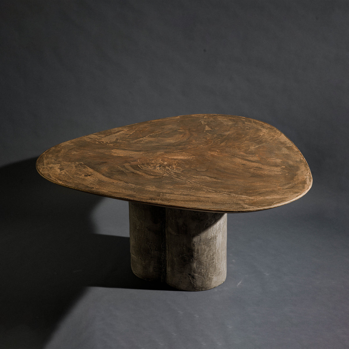 A handmade triangle-shaped dinning table with a bronze top and a triple round column base with dark gray stone texture, set against a dark gray studio background.