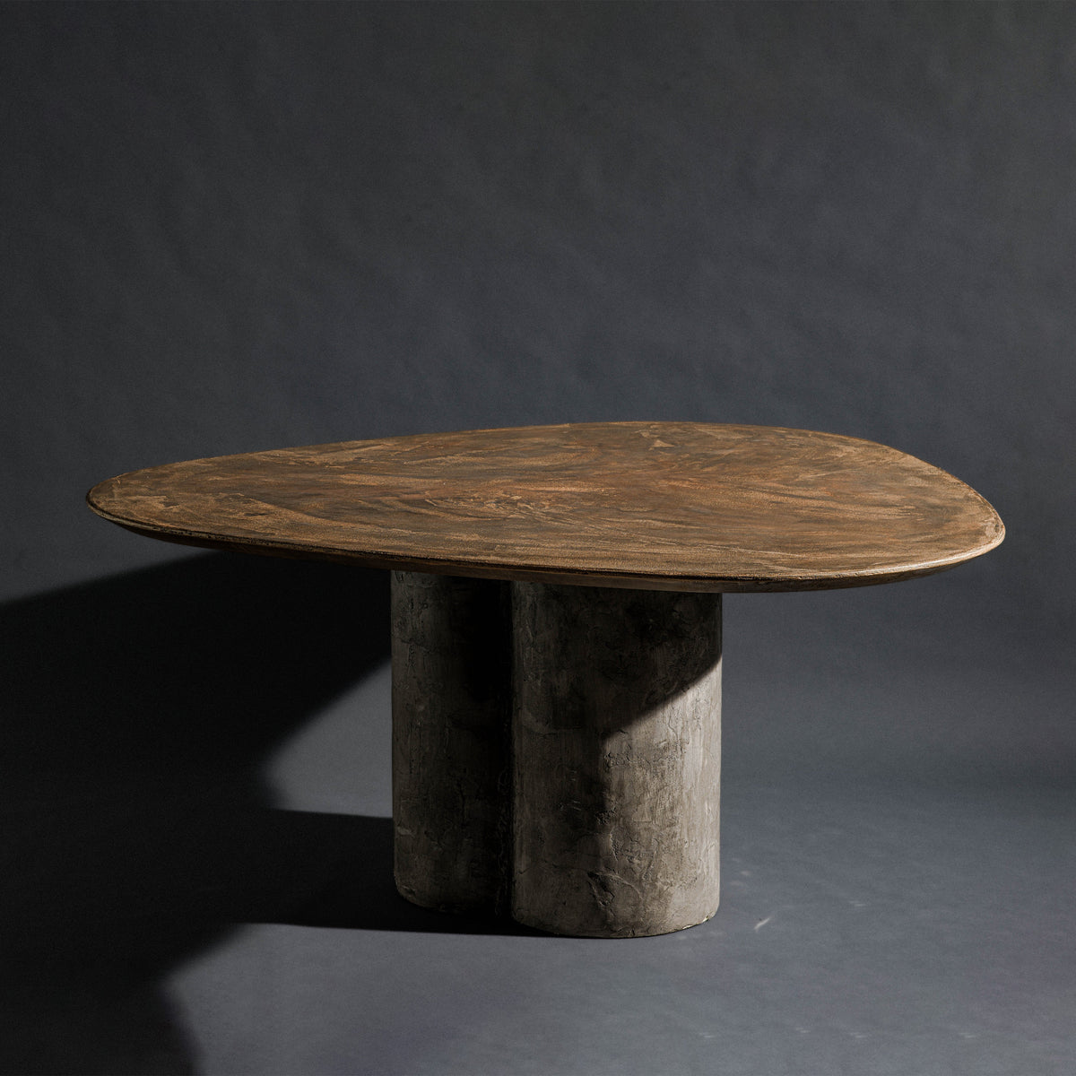 A handmade triangle-shaped dinning table with a bronze top and a triple round column base with dark gray stone texture, set against a dark gray studio background.