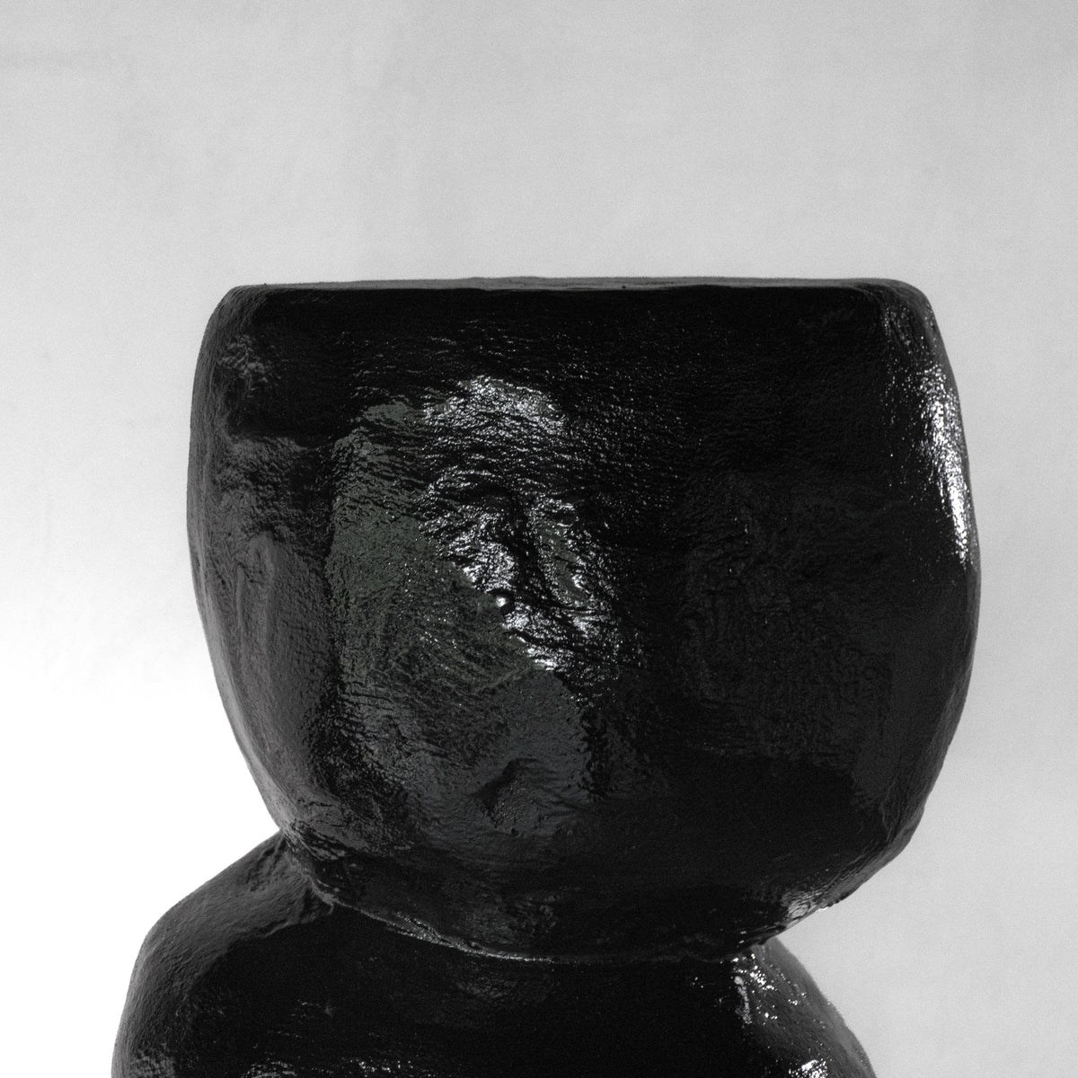 A close-up of a sculptural shiny black stool with a texture that has an organic shape, displayed on a white pedestal against a light gray background.