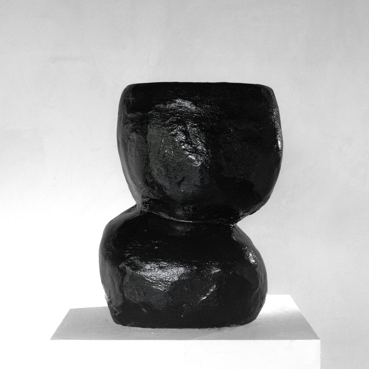 A sculptural shiny black stool with a texture that has an organic shape, displayed on a white pedestal against a light gray background.