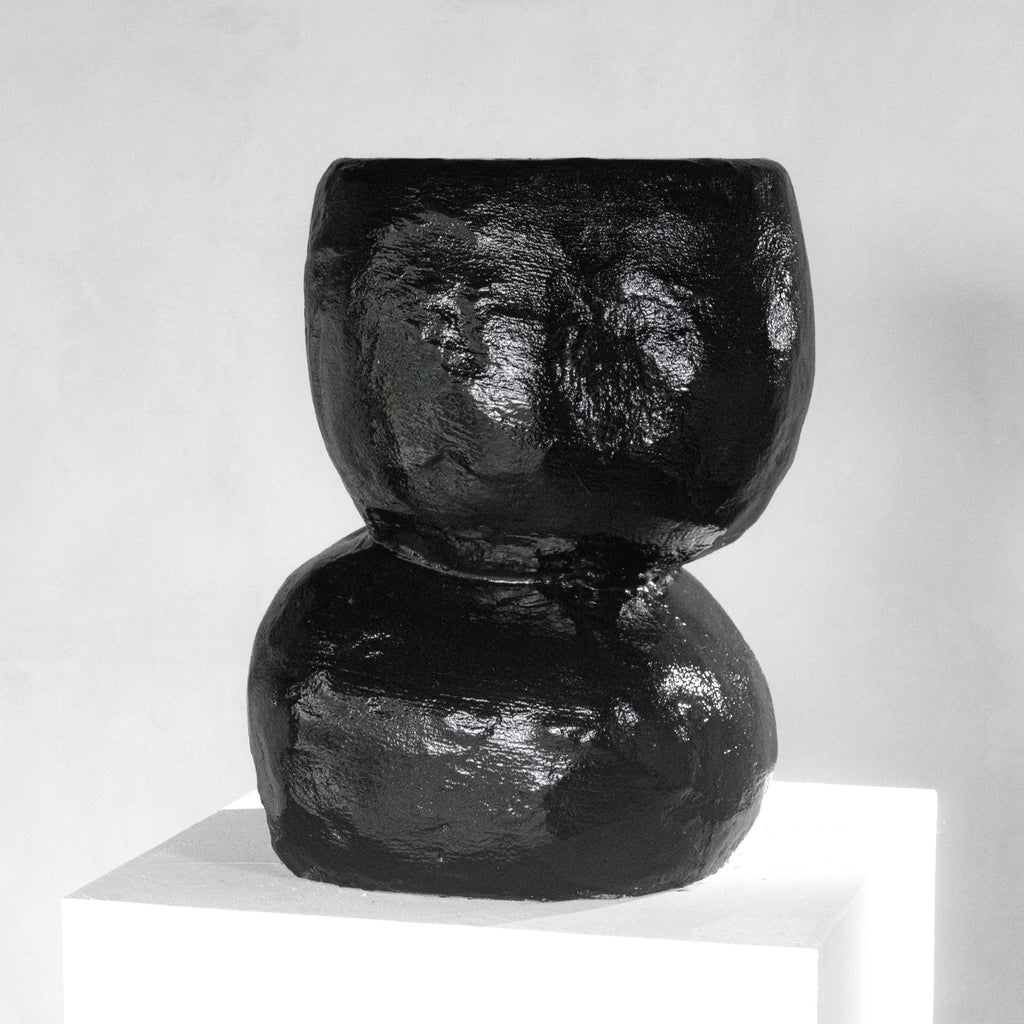 A sculptural shiny black stool with a texture that has an organic shape, displayed on a white pedestal against a light gray background.