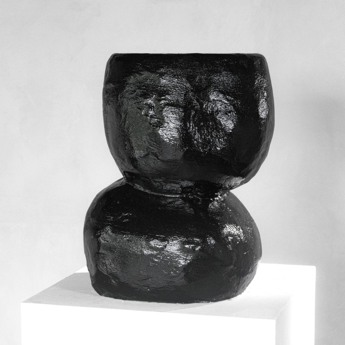 A sculptural shiny black stool with a texture that has an organic shape, displayed on a white pedestal against a light gray background.