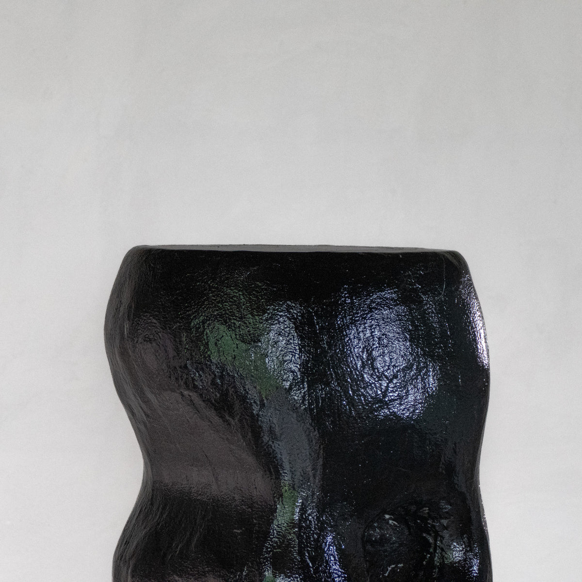 Close-up of an abstract sculptural black shiny stool with a hole, displayed on a white pedestal against a light gray background.