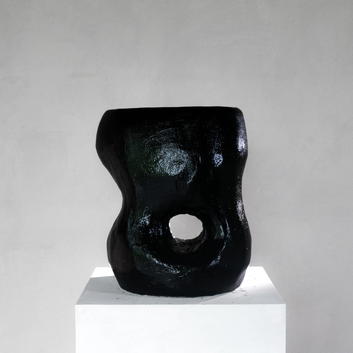 An abstract sculptural black shiny stool with a hole, displayed on a white pedestal against a light gray background.