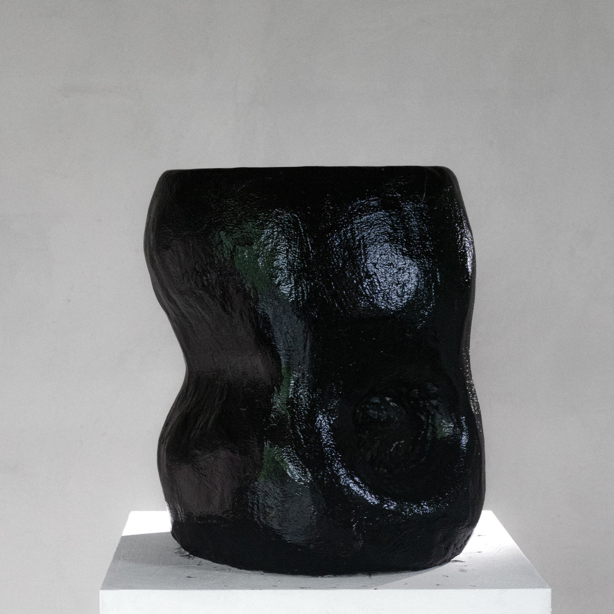 An abstract sculptural black shiny stool with a hole, displayed on a white pedestal against a light gray background.