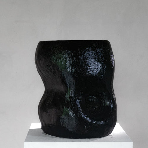 An abstract sculptural black shiny stool with a hole, displayed on a white pedestal against a light gray background.