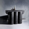 A handmade sculptural closet made of wood and finished with charcoal black texture, featuring three vertical cylindrical columns supporting a wide rectangular top, with two thin bars across the cabinet extending from the sides, set against a gray studio background.