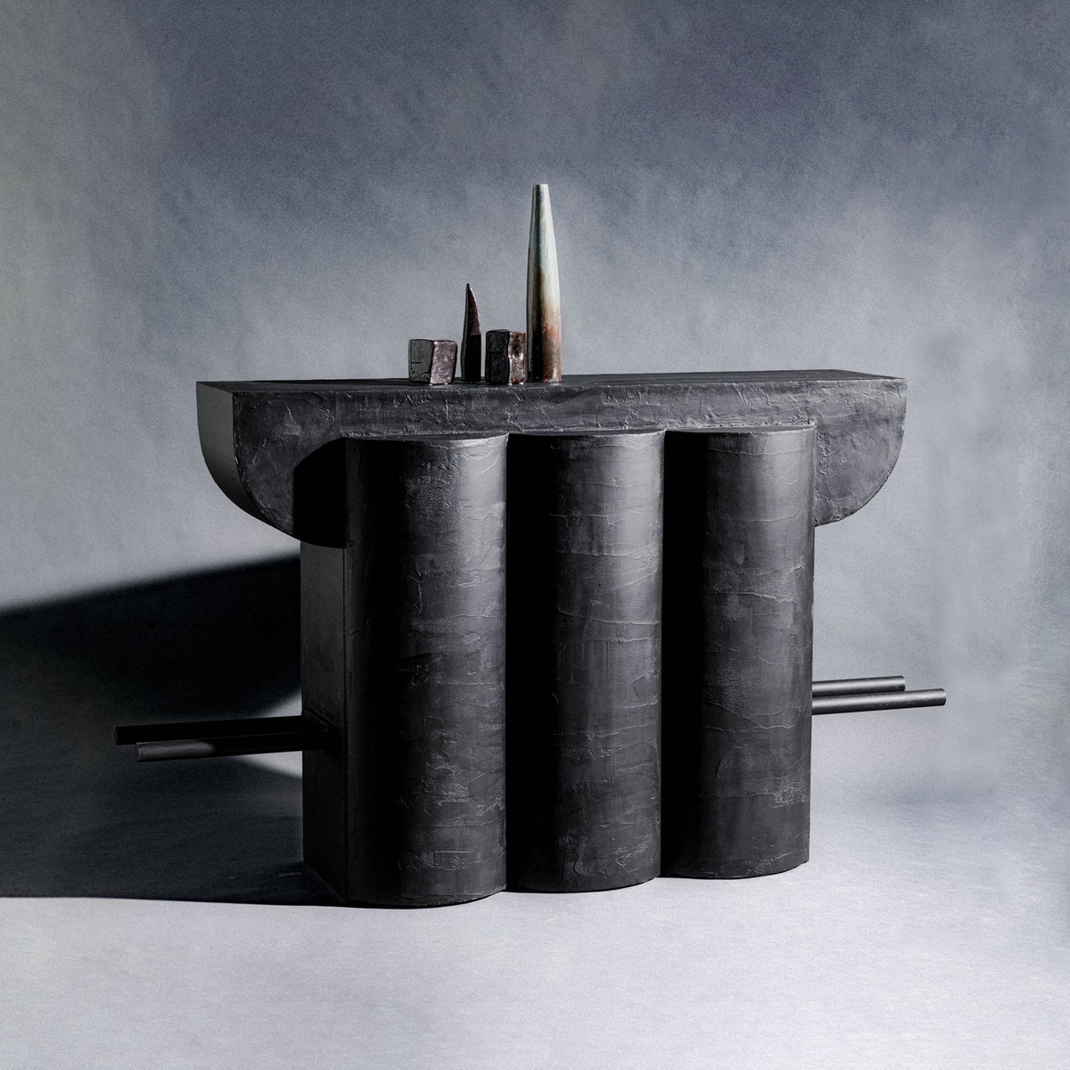 A handmade sculptural closet made of wood and finished with charcoal black texture, featuring three vertical cylindrical columns supporting a wide rectangular top, with two thin bars across the cabinet extending from the sides, set against a gray studio background.