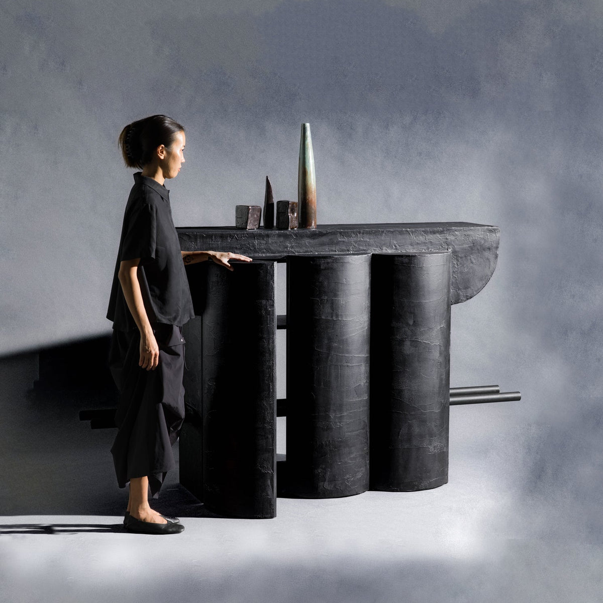 A handmade sculptural closet made of wood and finished with charcoal black texture, featuring three vertical cylindrical columns supporting a wide rectangular top, with two thin bars across the cabinet extending from the sides, set against a gray studio background.