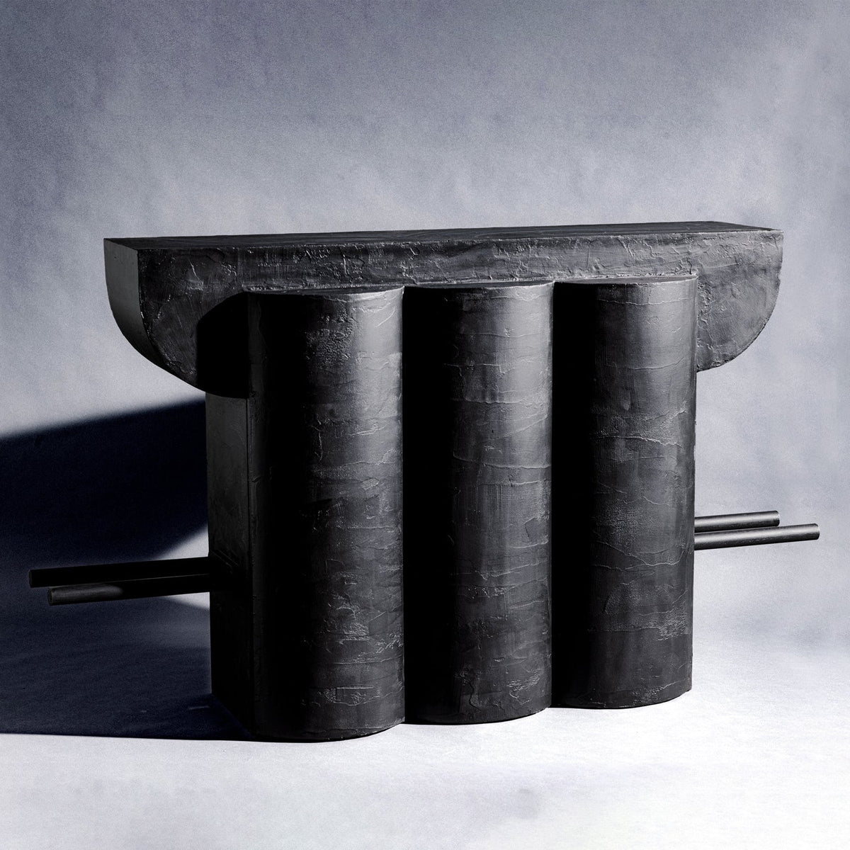 A handmade sculptural closet made of wood and finished with charcoal black texture, featuring three vertical cylindrical columns supporting a wide rectangular top, with two thin bars across the cabinet extending from the sides, set against a gray studio background.