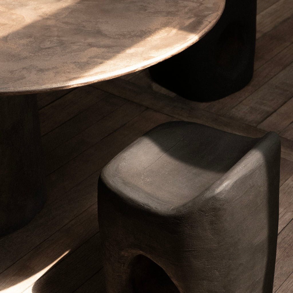 A close-up of a sculptural dark gray concrete stool in a sunlit room, with a matching round table and a black stool in the background on a wood floor.
