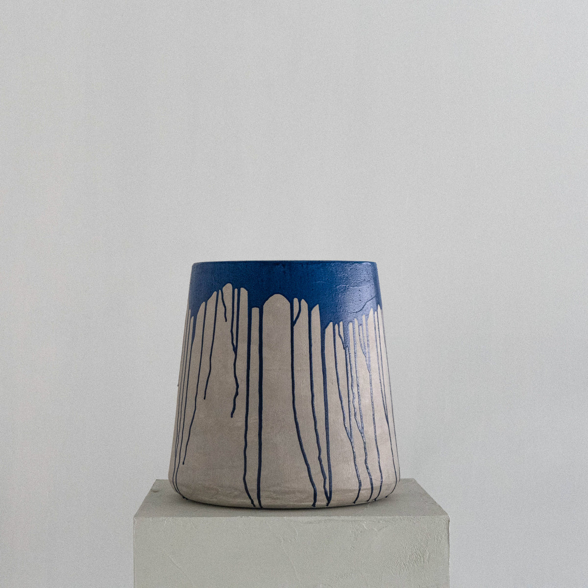 A sculptural stool, light grey body with a bright blue top and dripping down over the grey base, stands on pedestal against a light gray background.