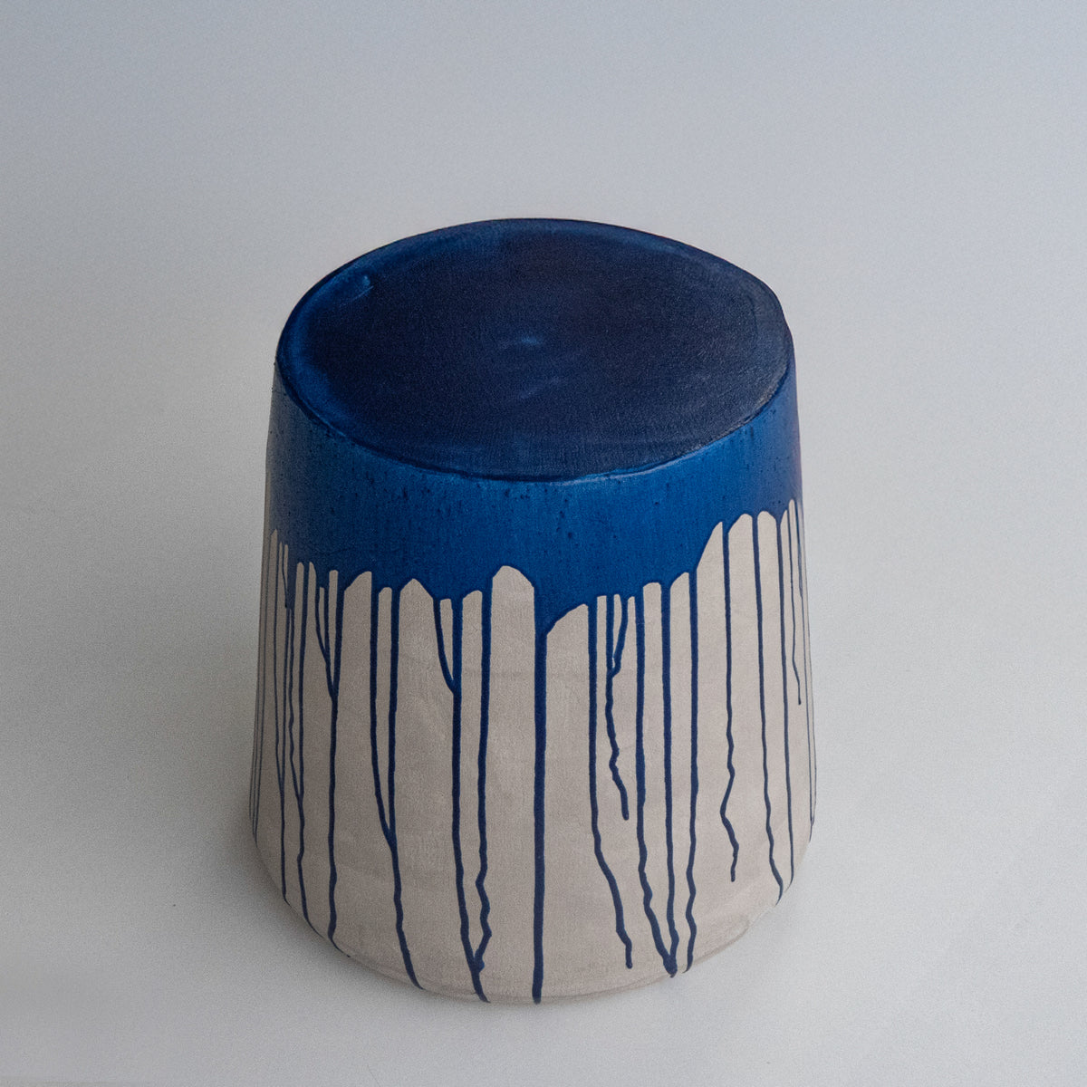 A sculptural stool, light grey body with a bright blue top and dripping down over the grey base, stands against a light gray background.