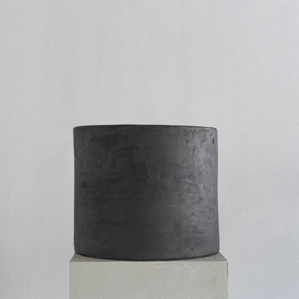 A sculptural stool finished in micro-cement and limestone. Sculpted into an abstract shape that features a curved, copper-toned metal half-shell resting over a matte, dark grey rectangular block. 