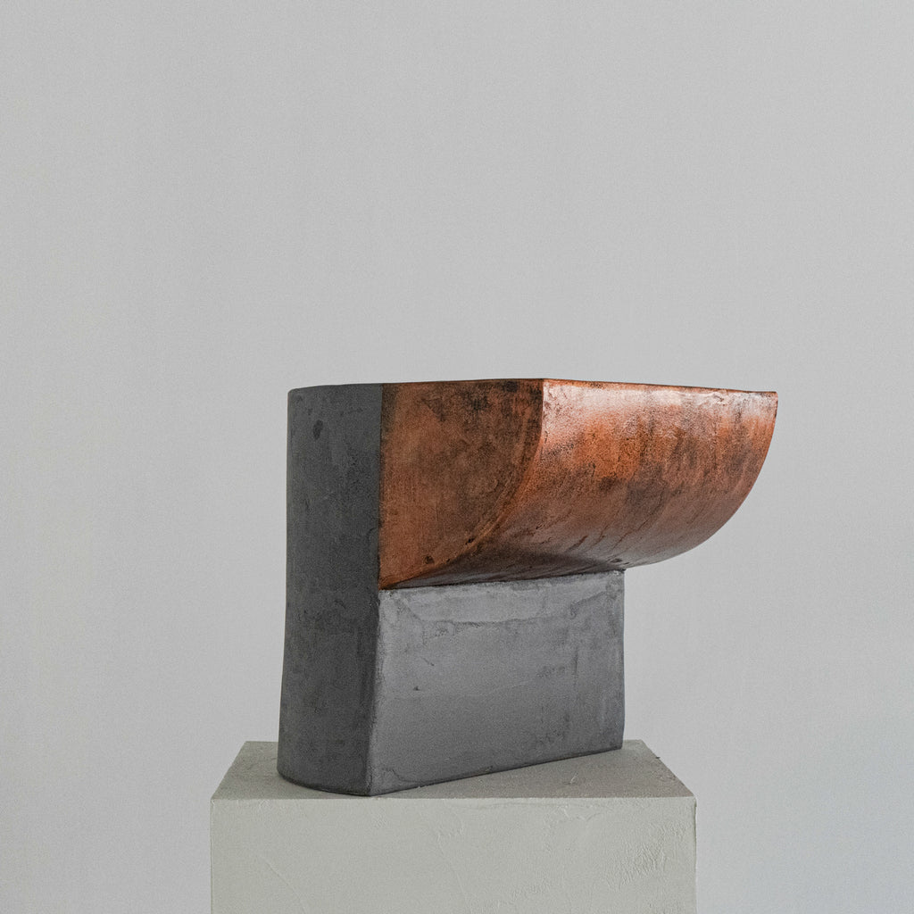 A sculptural stool finished in micro-cement and limestone. Sculpted into an abstract shape that features a curved, copper-toned metal half-shell resting over a matte, dark grey rectangular block. 