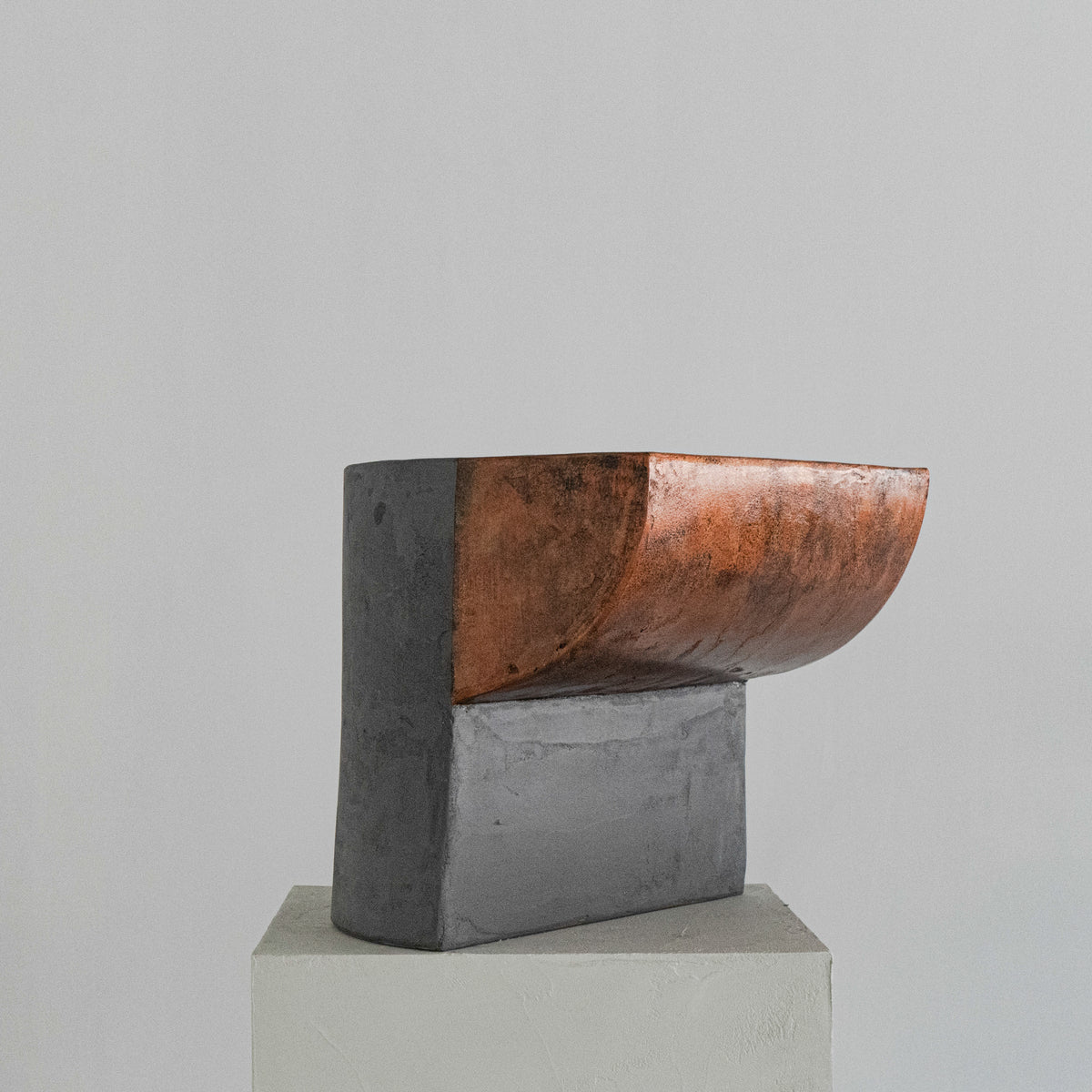 A sculptural stool finished in micro-cement and limestone. Sculpted into an abstract shape that features a curved, copper-toned metal half-shell resting over a matte, dark grey rectangular block. 