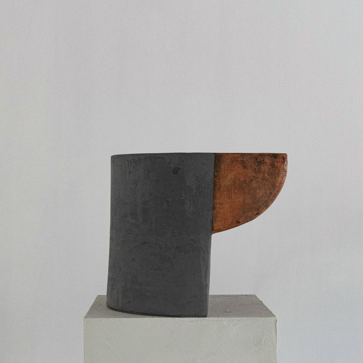 A sculptural stool finished in micro-cement and limestone. Sculpted into an abstract shape that features a curved, copper-toned metal half-shell resting over a matte, dark grey rectangular block. 