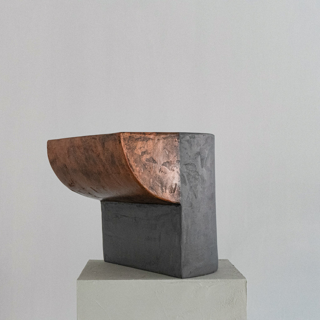 A sculptural stool finished in micro-cement and limestone. Sculpted into an abstract shape that features a curved, copper-toned metal half-shell resting over a matte, dark grey rectangular block. 