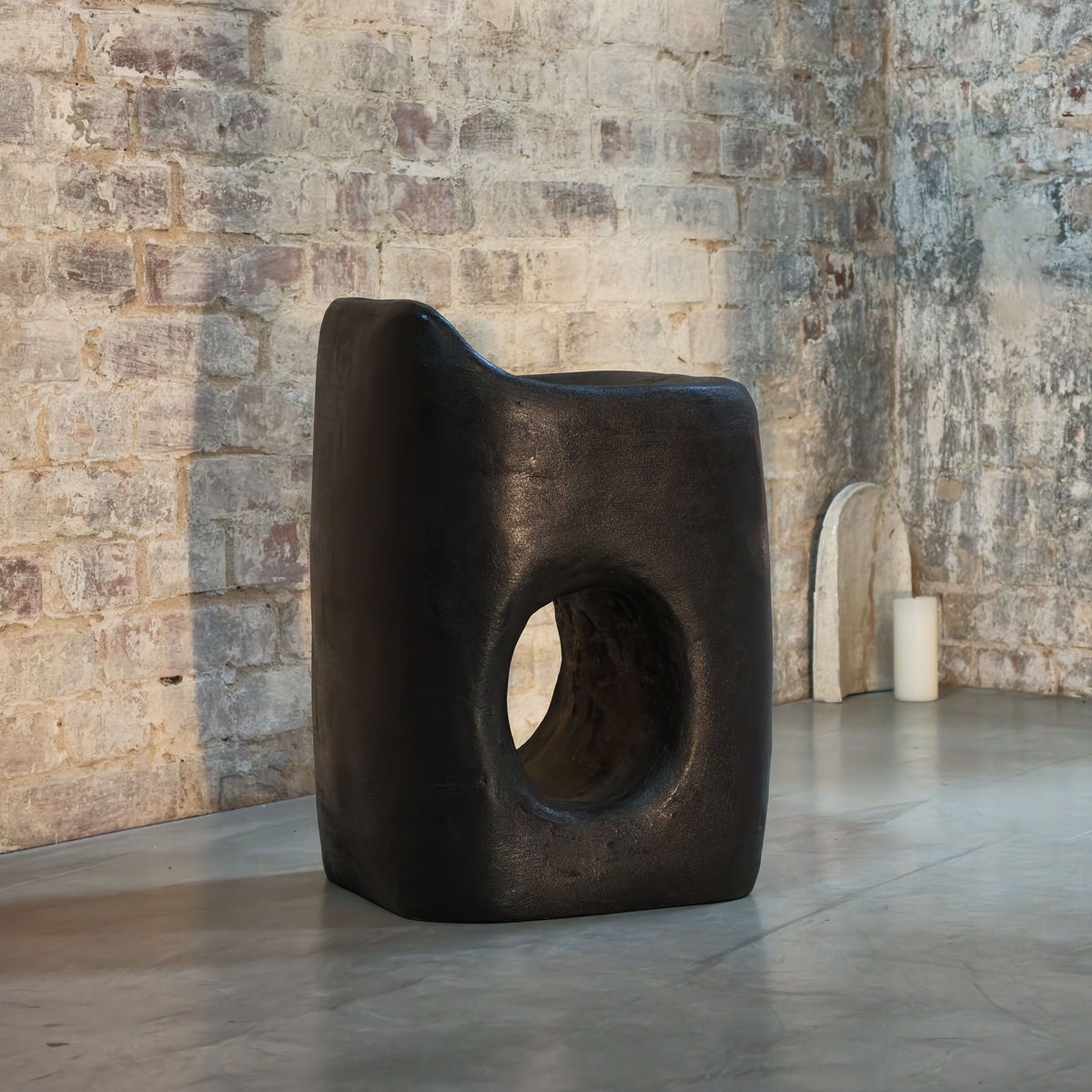 A sculptural black stone chair with a large circular cutout in the center, set in a gallery space with a rustic brick wall and concrete floor.